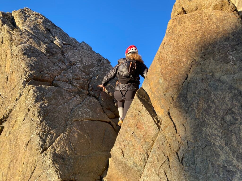 A woman with a red climbing helmet and black backpack climbing through two large rocks. 