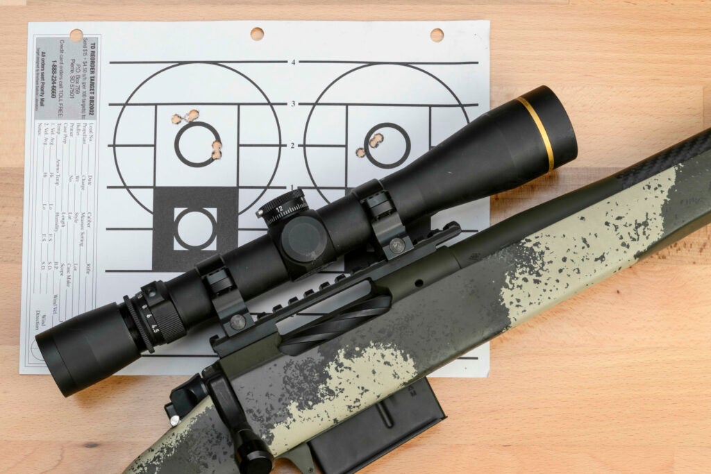 A camo-stocked bolt-action rifle, topped with a black scope, lying on top of a paper target with two zeroing groups.
