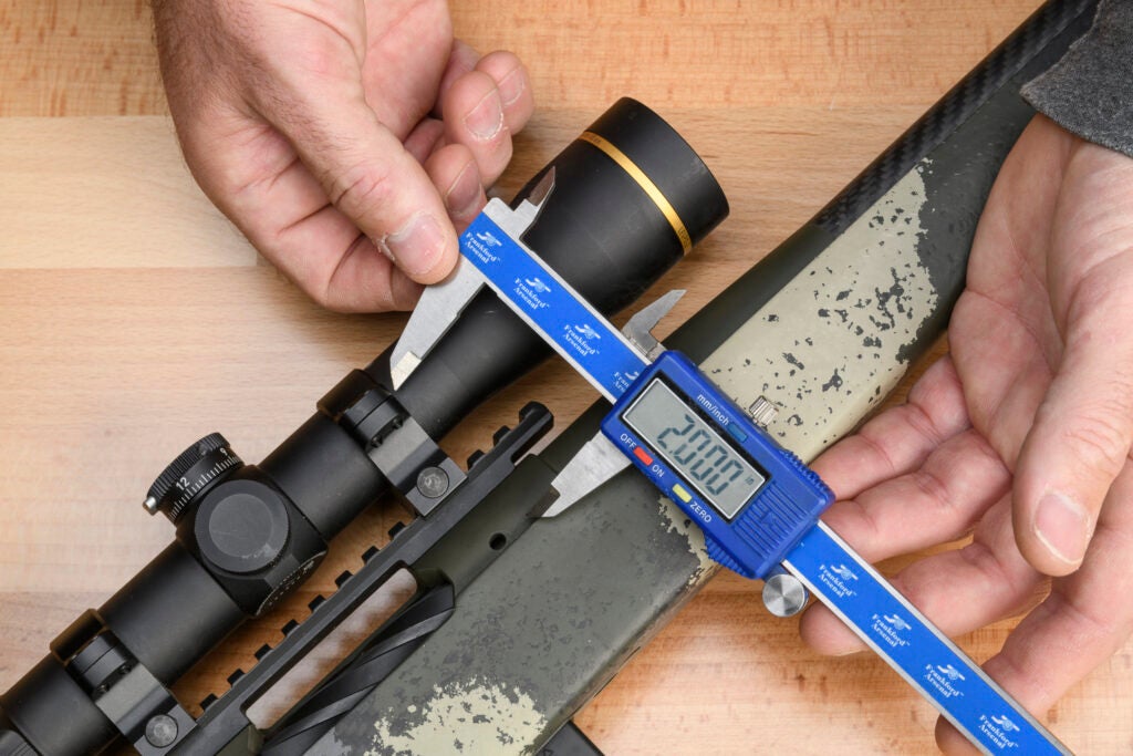A pair of hands uses a blue digital caliper to measure the center of the rifle bore to the center of the scope.