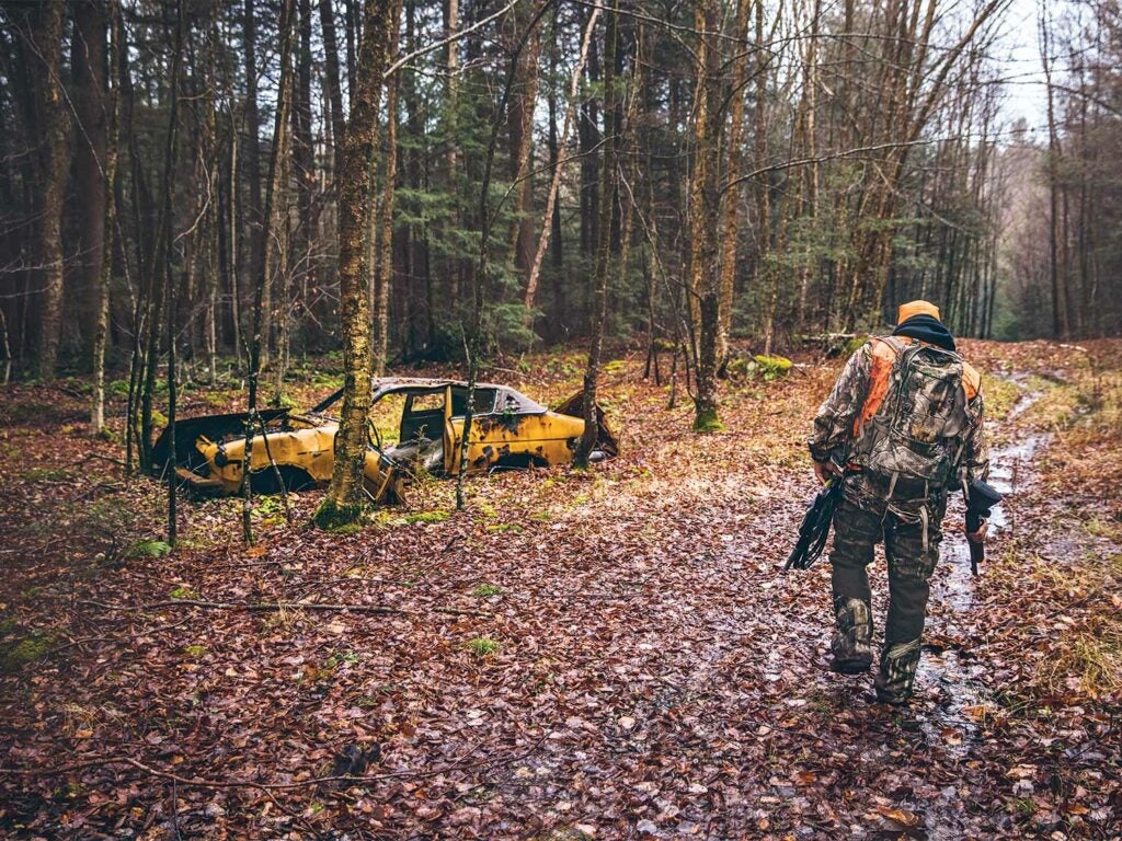 hunter walking through the woods with an abandoned rusty car nearby