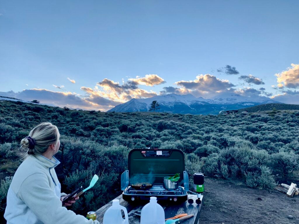 A woman cooking on a green camp stove overlooking a mountain