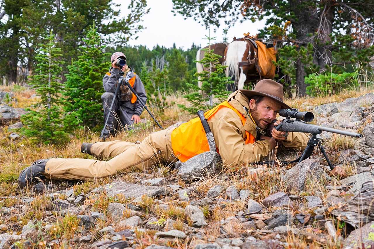 hunter looks through scope, with spotter and horses behind him