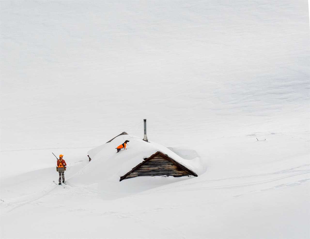 A hunter and her bird dog ski near a snowed-in mining building. 