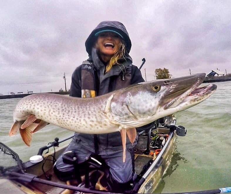 A woman in a jacket holding a giany musky on the water