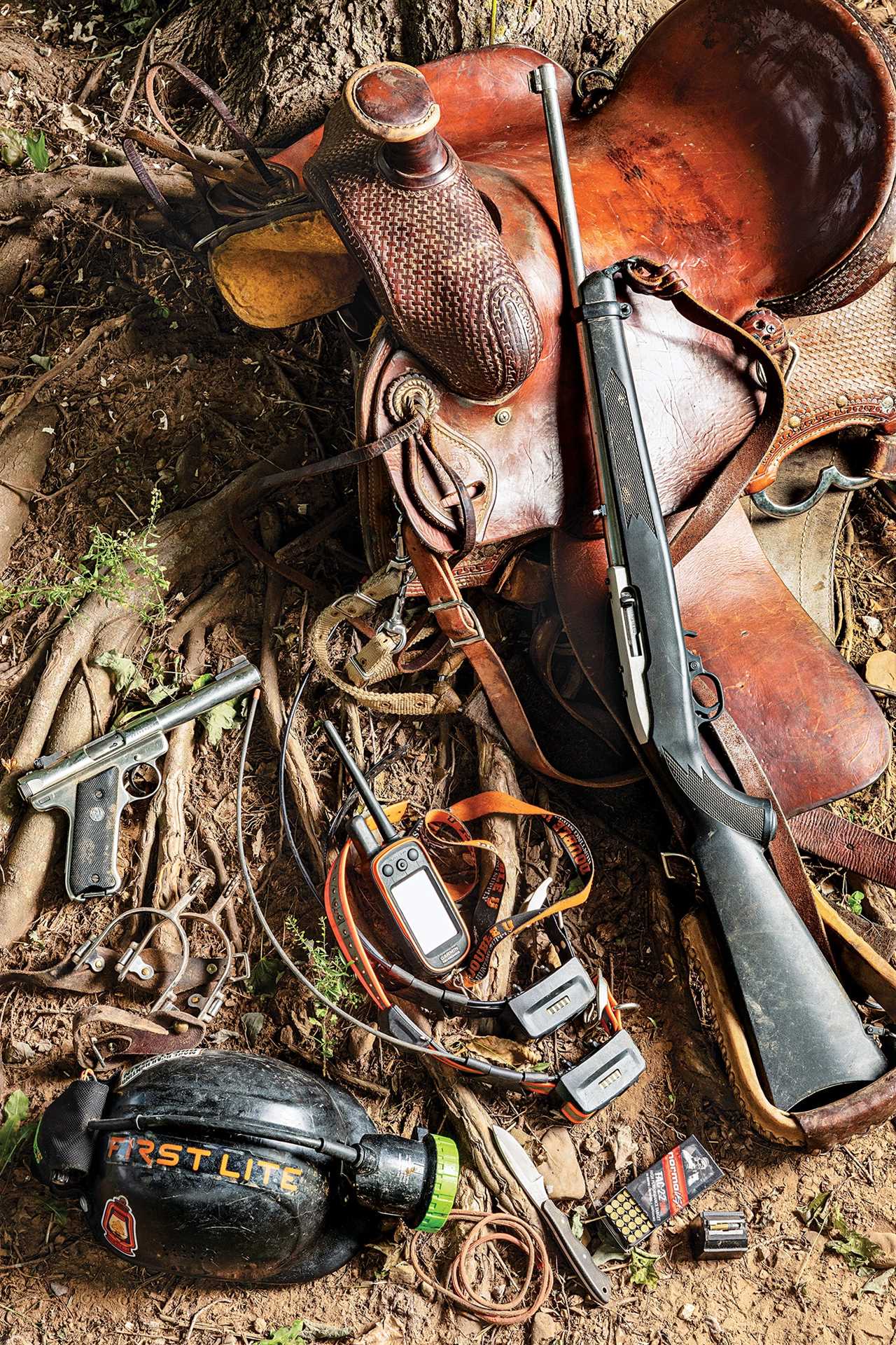 saddle with other raccoon hunting equipment: a rifle, a handgun, a gps unit, a helmet with a headlamp, tracking collars, and more