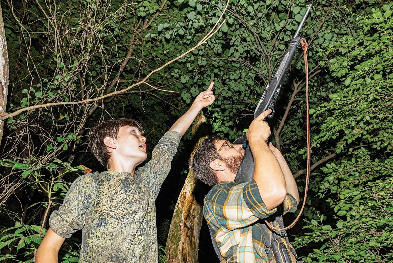 boy points up into tree while man aims rifle into the foliage