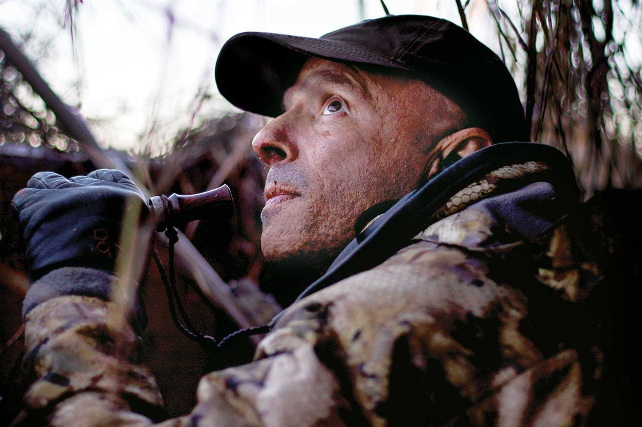 hunter peers up, holding duck call in gloved hand, ready to use it
