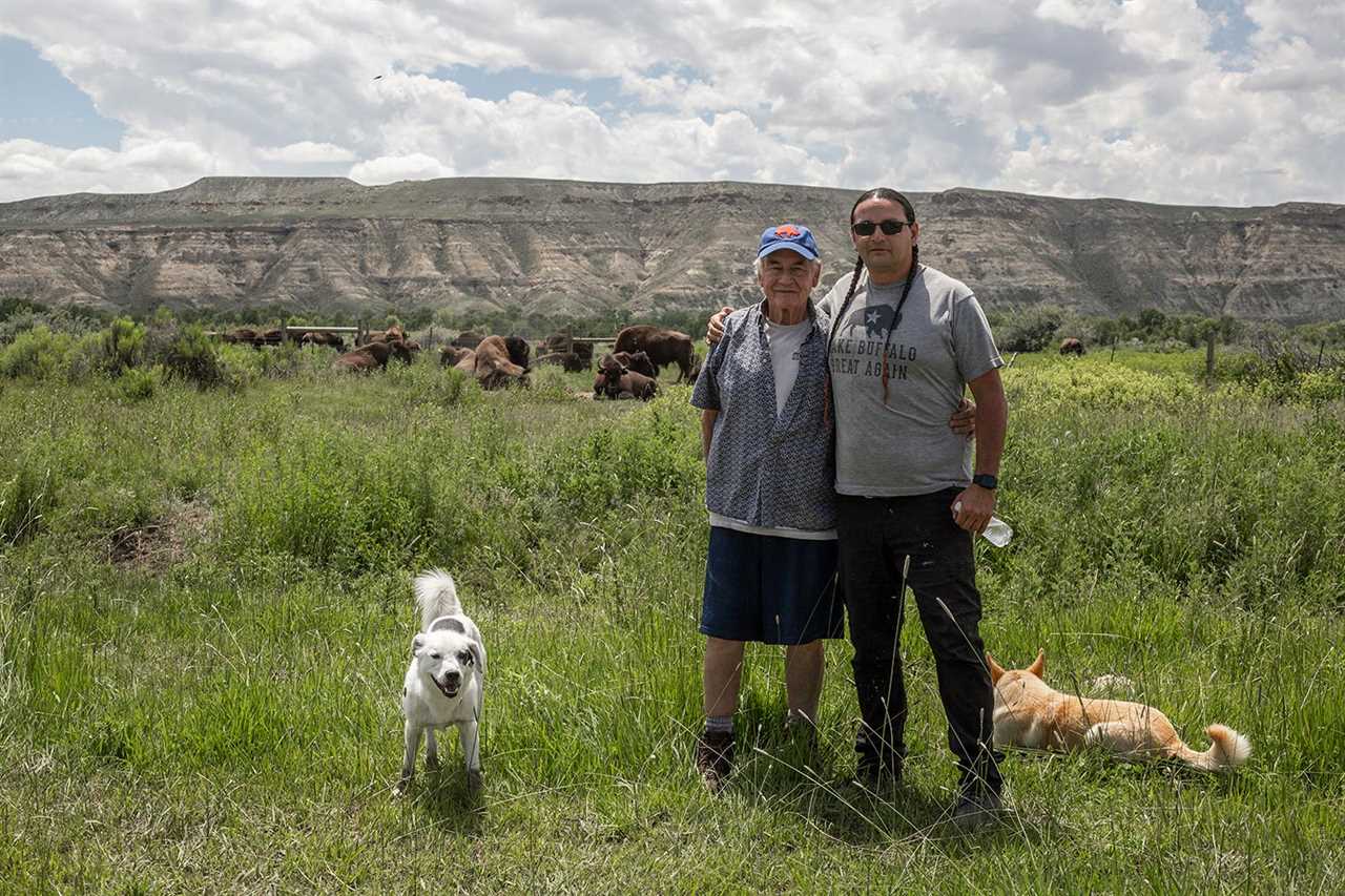 two men and stand on prairie with two dogs; buffalo and cliffs behind them