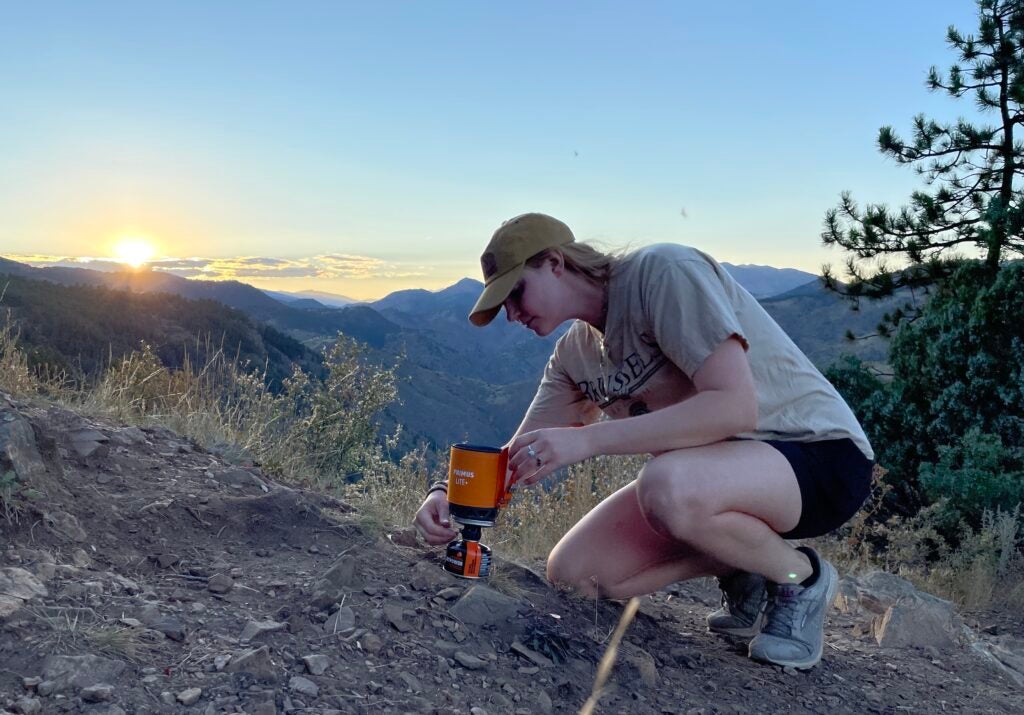 A woman setting up an orange camping stove in front of mountains