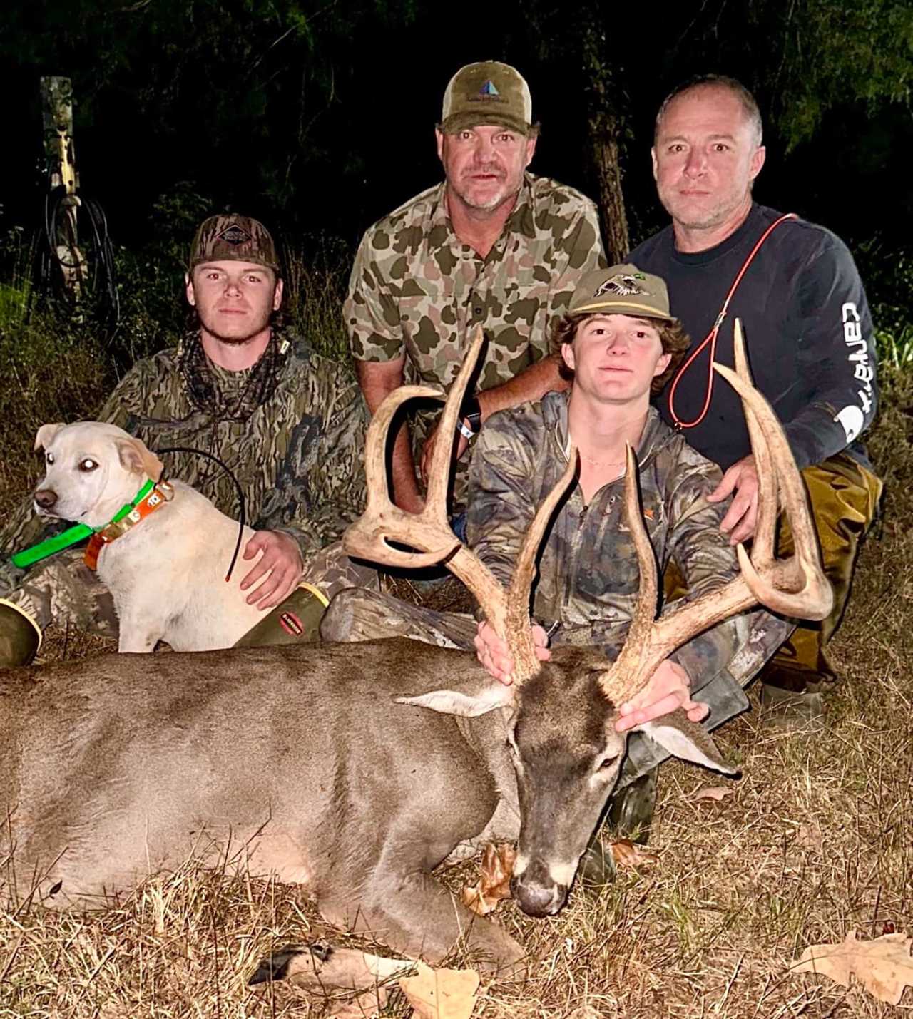 Hunters and a tracking dog sit behind a deer.